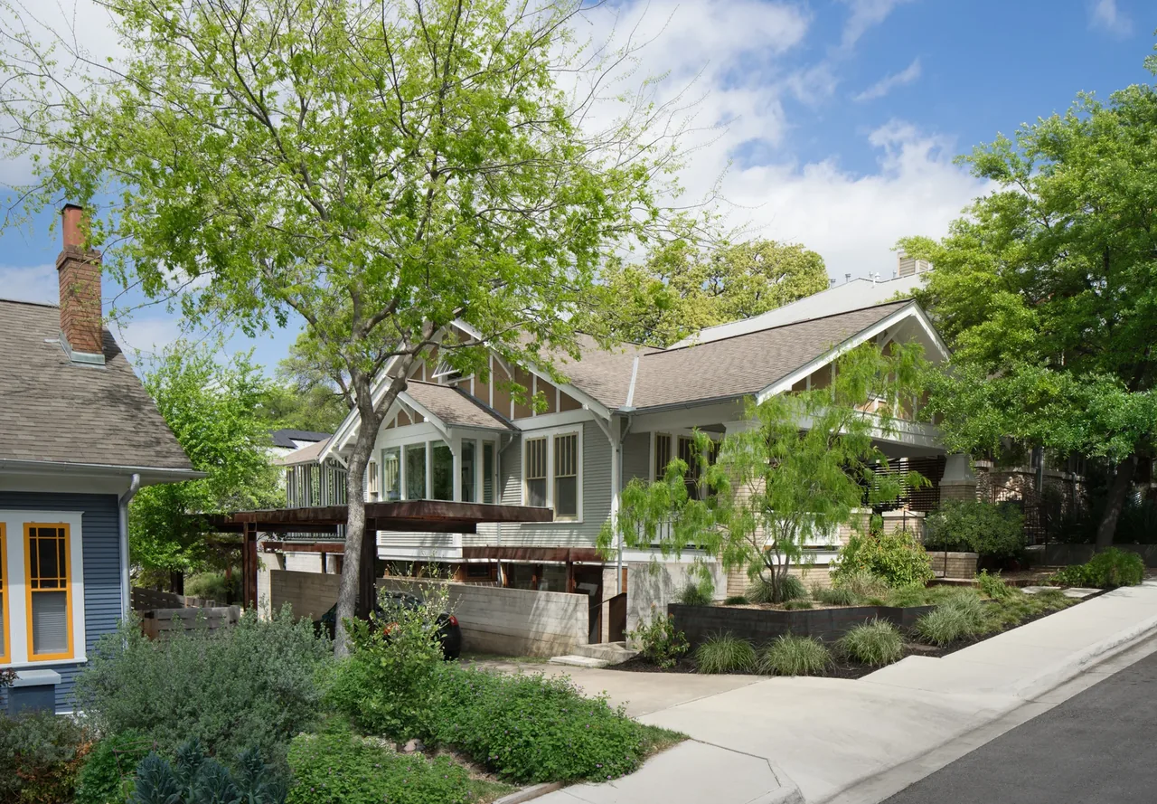 A house features a gable roof, light gray siding, and a raised porch entry amidst mature landscaping and a retaining wall.