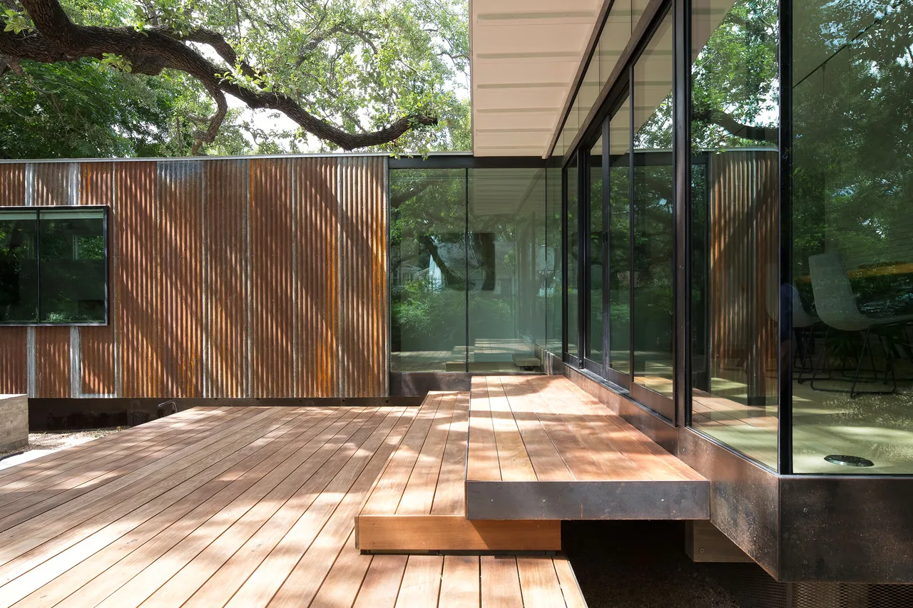 Wood deck with built-in wooden bench and large floor-to-ceiling glass windows with black frames beside a rusted metal siding wall.