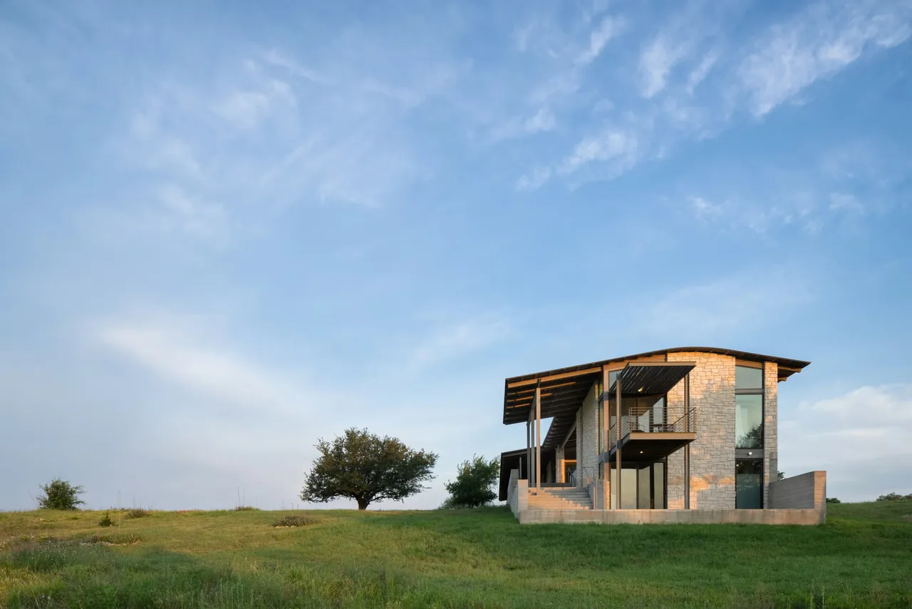 Modern two-story house with stone siding and large overhanging roof on a grassy hill.