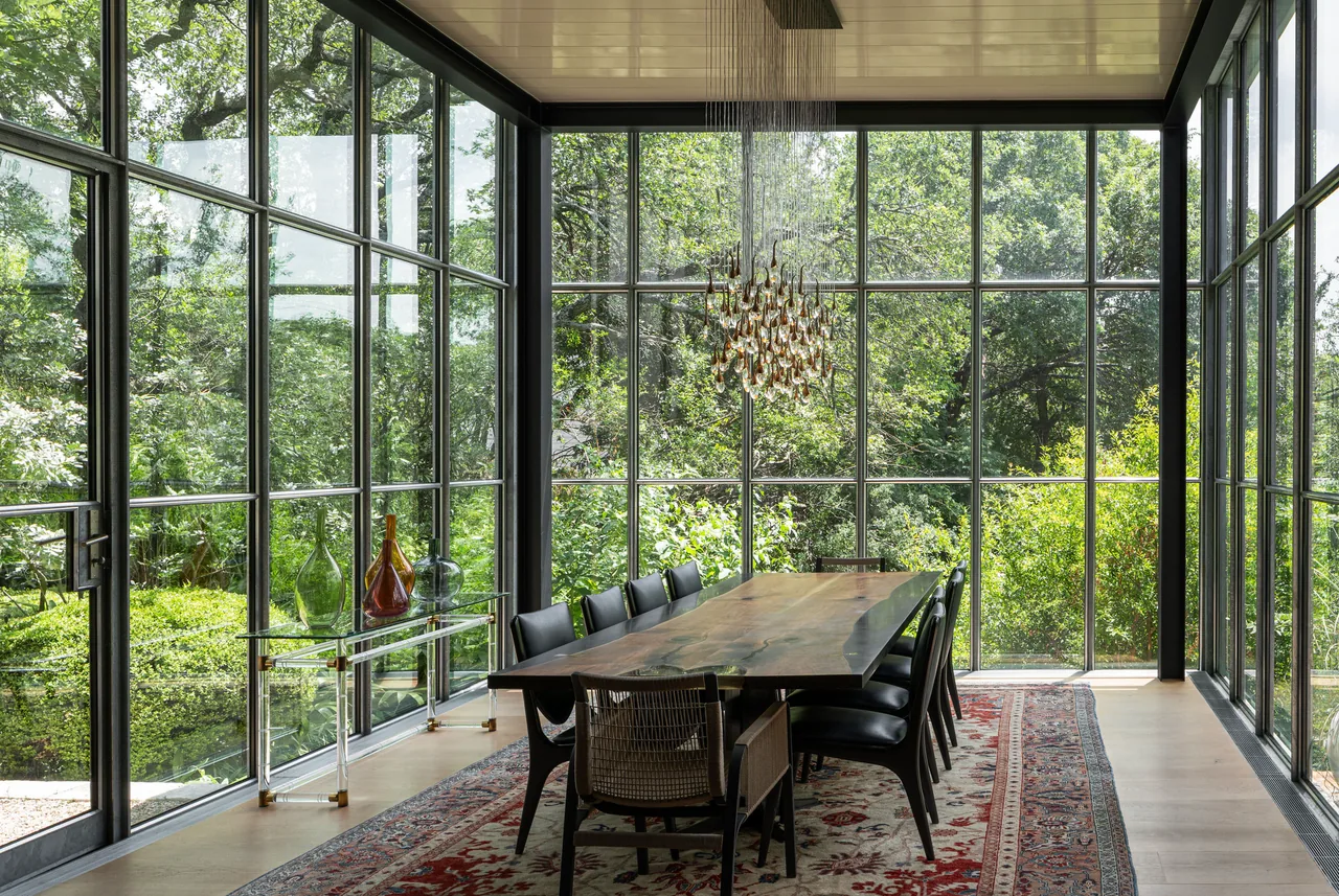 Dining room features floor-to-ceiling black-framed glass walls with views to greenery and a wood dining table under a sculptural chandelier.
