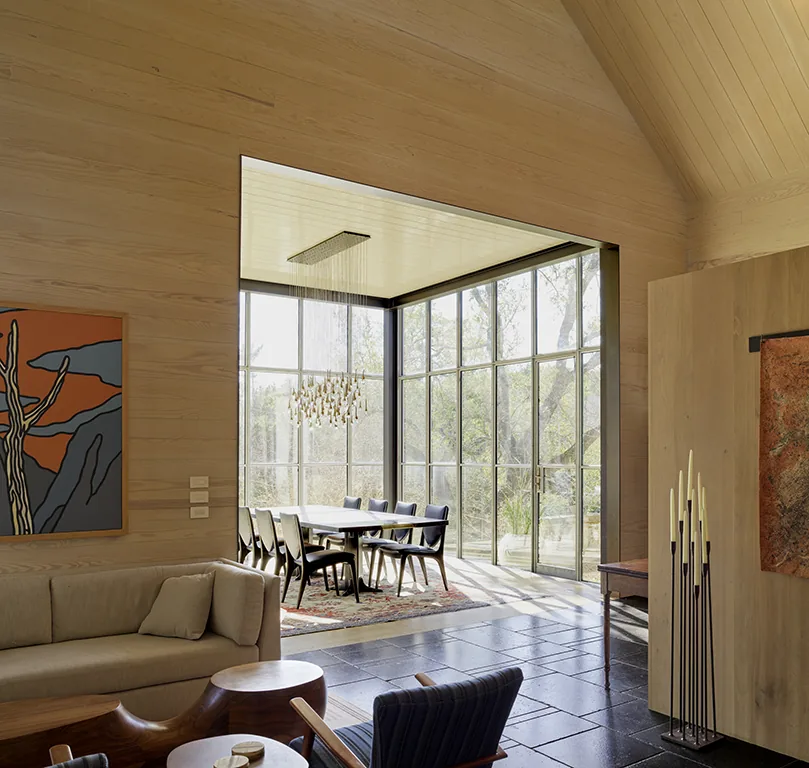 Dining area with large grid-format glass windows, wooden-paneled walls and ceiling, and a wood slab table under a sculptural chandelier.