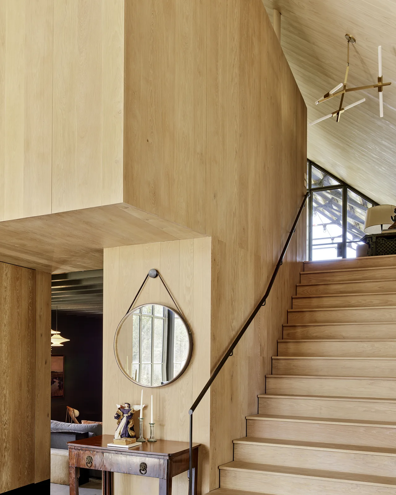Interior wood-paneled stair with a round mirror over a wooden console table and light fixture above.