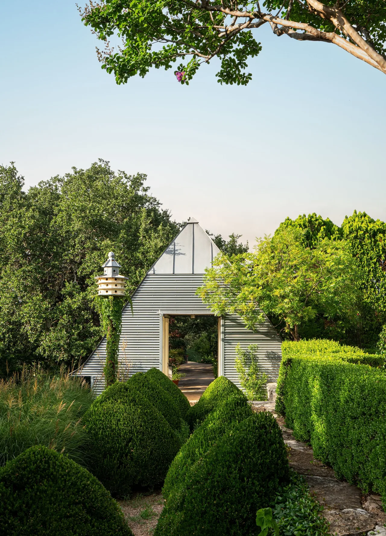 Triangular corrugated metal garden structure with open doorway surrounded by shaped green hedges and trees.