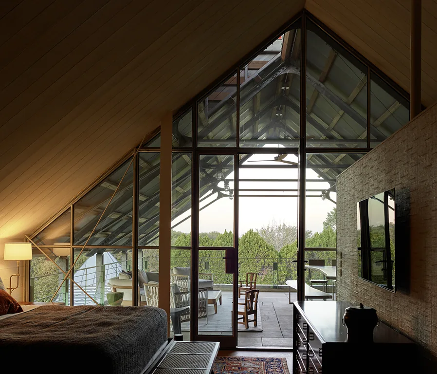 Interior view of a bedroom with a vaulted wood ceiling and floor-to-ceiling glazing to a covered patio with seating and nature beyond.