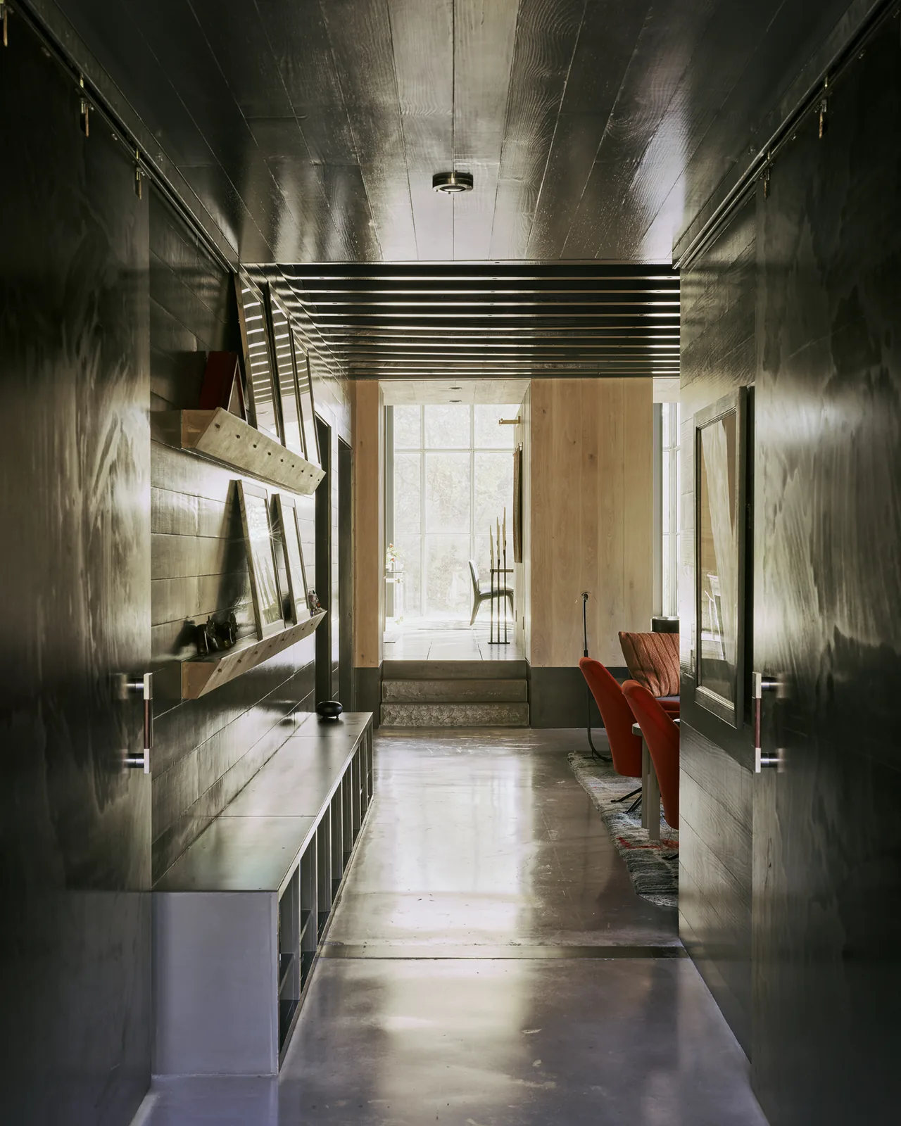 Contemporary dark hallway with black wood-paneled walls, concrete flooring, and shelving leading to a bright adjacent room with red chairs.