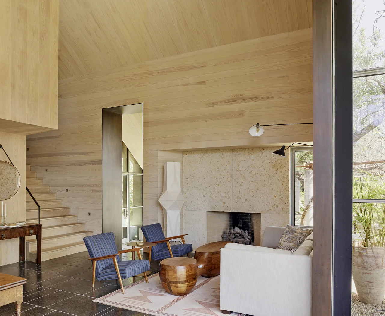 Living room with sloped wooden ceiling, stone fireplace, blue armchairs, and light-colored sofa overlooking garden.