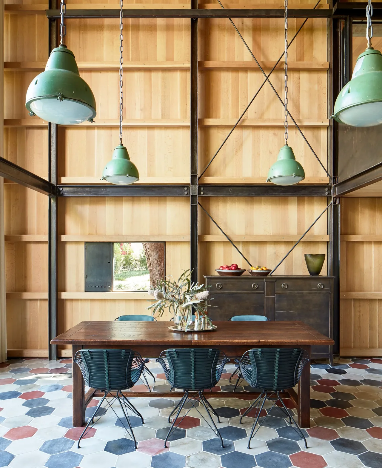Dining area featuring a hexagonal tiled floor, wire dining chairs, and industrial green pendant lights in a wood-paneled and steel-framed space.