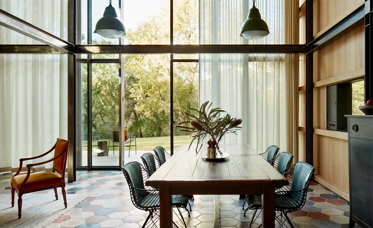 The Jewell Box dining space features a rustic wooden table paired with wire chairs and large glass walls facing a natural landscape in Austin.