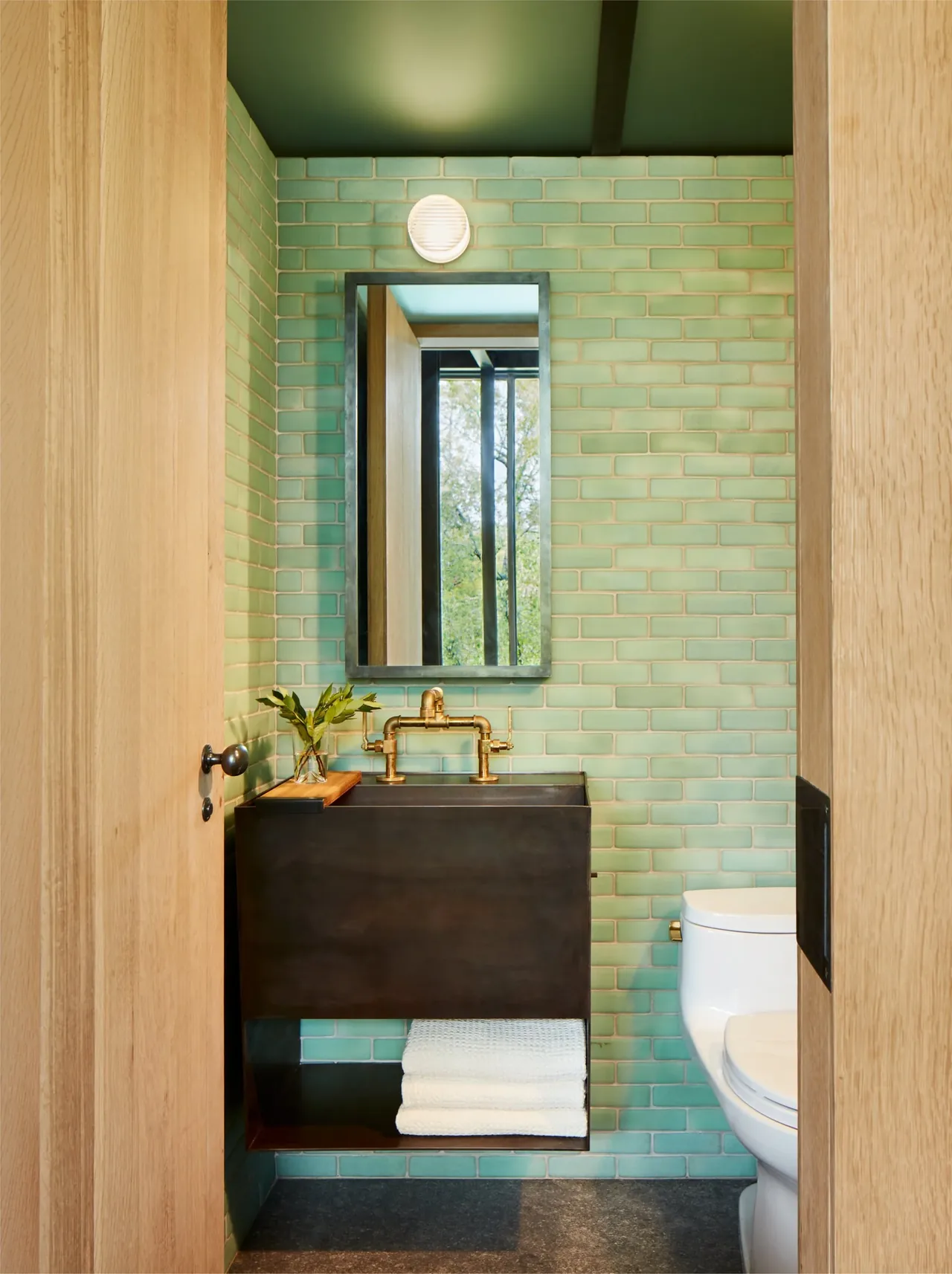 Modern bathroom with green subway tile walls, floating dark sink cabinet, brass faucet, and white toilet in Jewell Box project by Mell Lawrence Architects.
