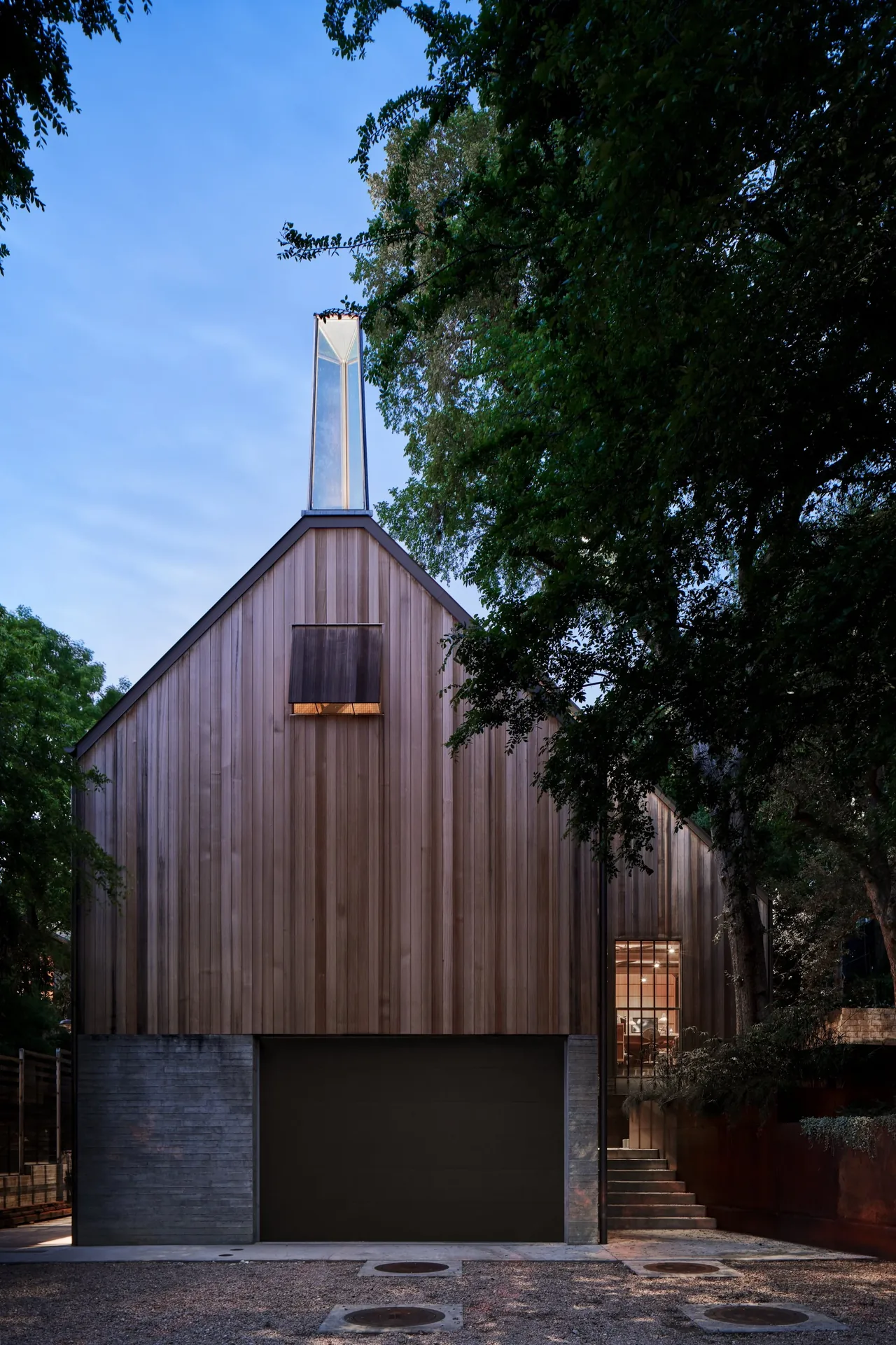 Modern wooden house features a tall glass chimney and concrete base, nestled among trees at dusk.