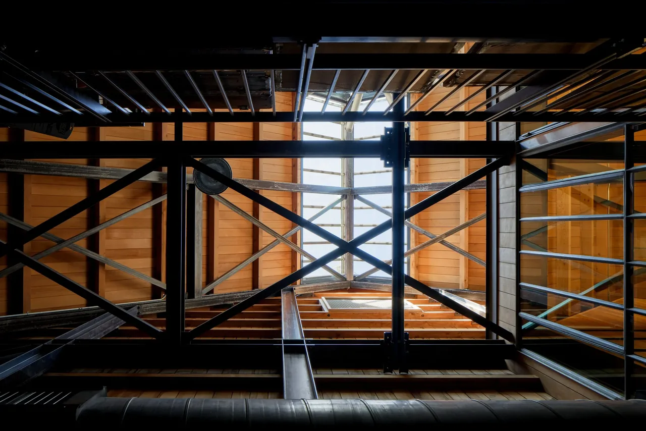 Interior view looking up through metal structural bracing to a wood-paneled ceiling with a skylight above.