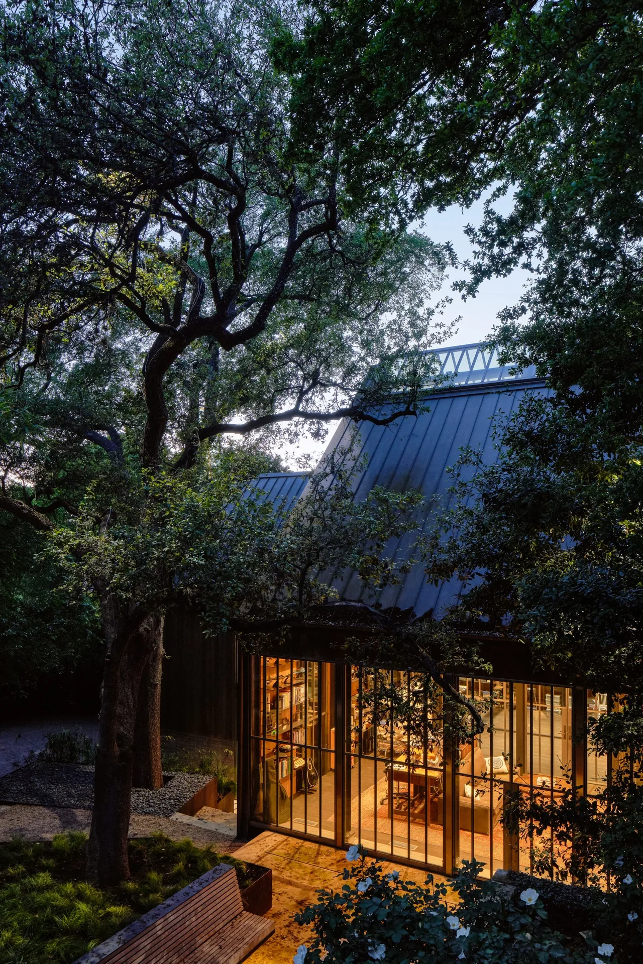 Evening exterior view of a house with black metal roof and large grid windows revealing a warmly lit interior library and living space.
