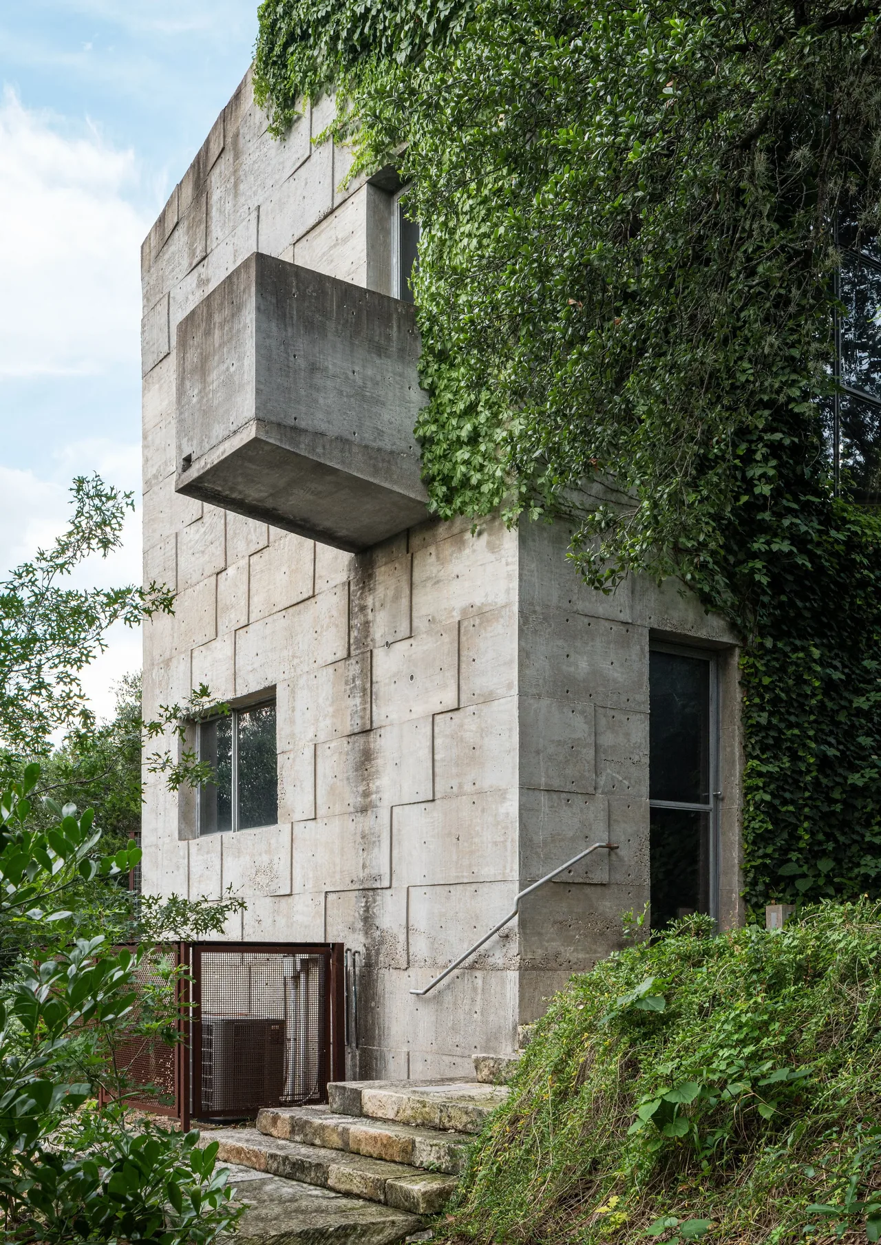 Concrete Studio Gallery by Mell Lawrence Architects features textured concrete walls with a small rectangular window and lush greenery.
