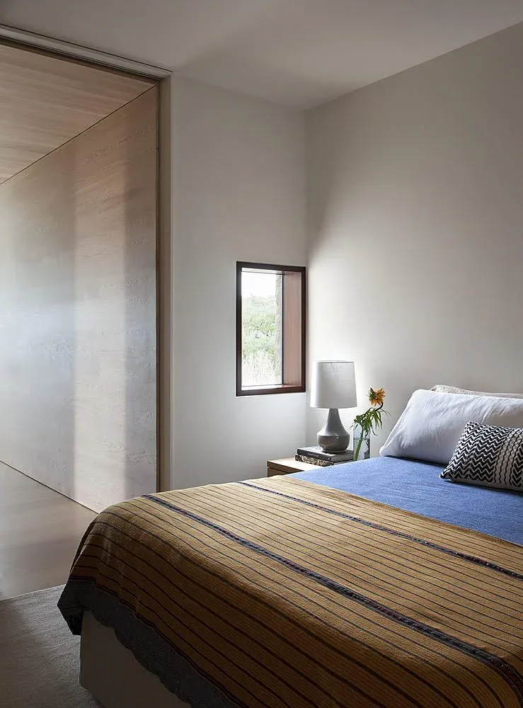 The 556 House bedroom features a small, framed window and a neutral color palette with simple furnishing.