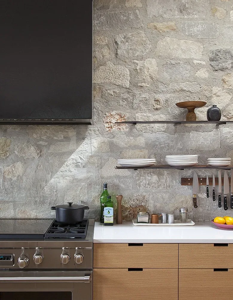 Kitchen with exposed stone backsplash, floating shelves holding plates and decor, and a stainless steel gas stove with a range hood.