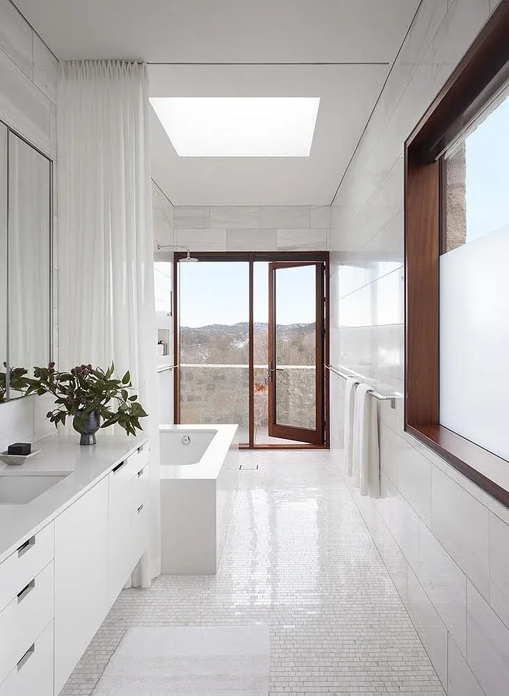 This modern bathroom features a freestanding tub, large wooden-framed window, and skylight with a view through glass doors.