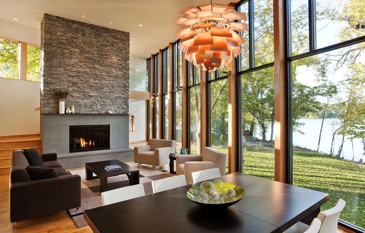 Living and dining space with a tall stone fireplace, large western-facing floor-to-ceiling windows, and modern furnishings.