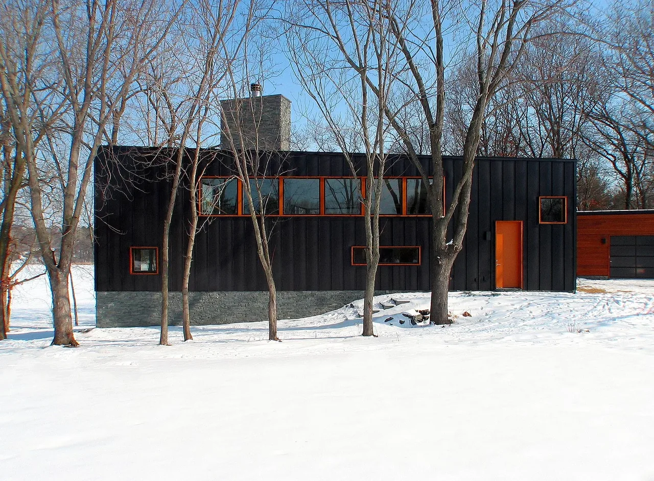 Farquar Lake Residence features a black exterior with stacked stone foundation and orange window frames in a snowy setting.