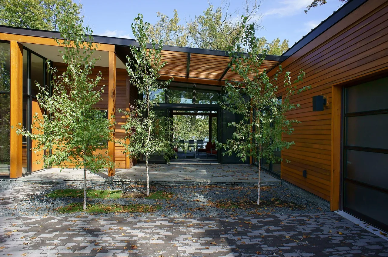The Farquar Lake Residence features wood siding, a glass door entry, and a paved driveway with scattered small trees in front.