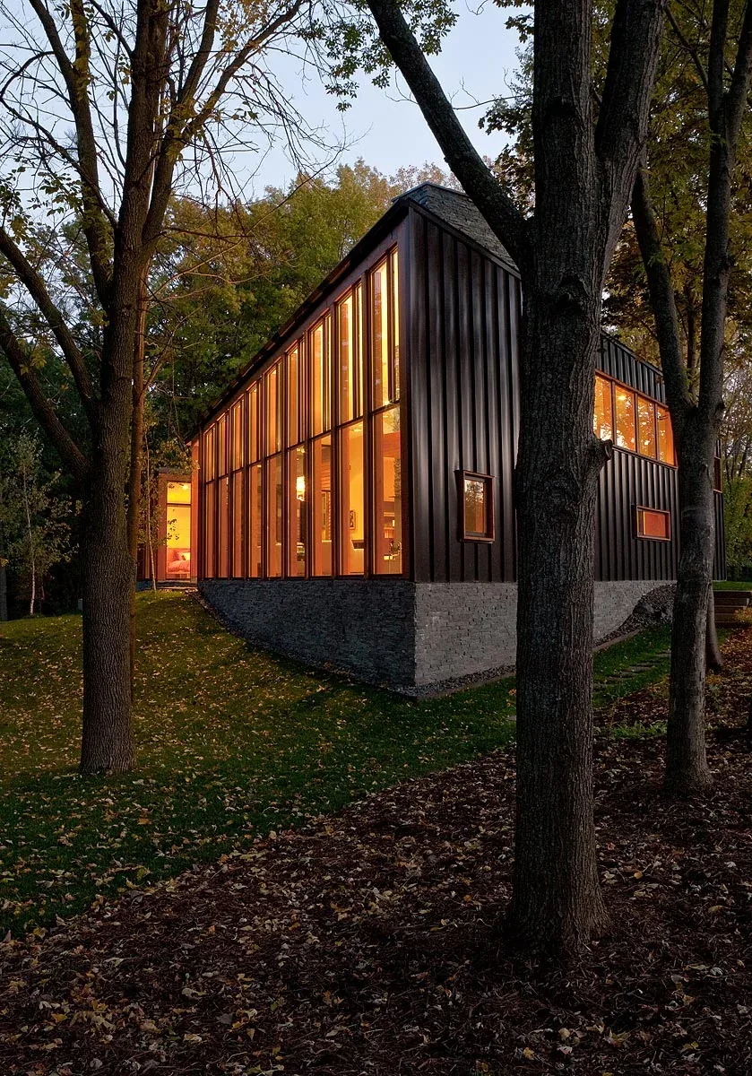 Modern residence with large vertical windows and stacked stone base, nestled among mature trees at dusk.