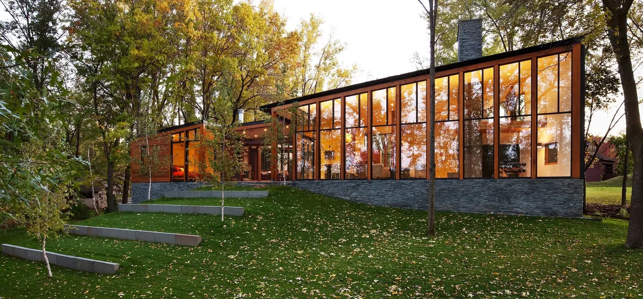 Farquar Lake Residence features floor-to-ceiling glazing on a stone foundation surrounded by trees and a sloping grassy yard.