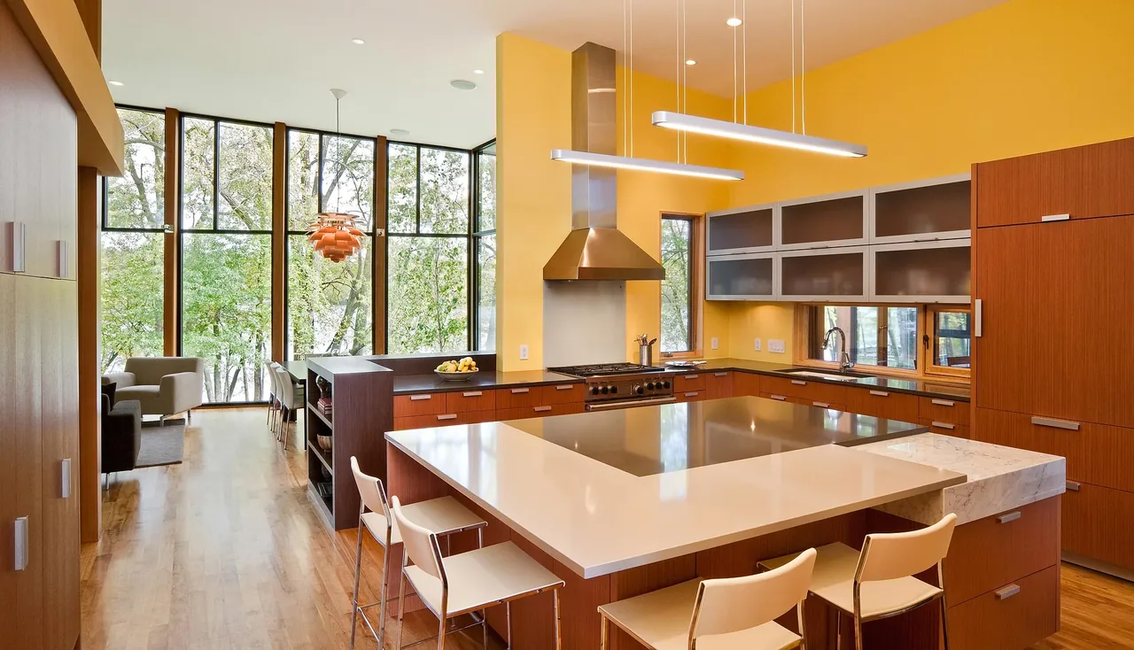 Open kitchen with wood cabinets, large island cooktop, and floor-to-ceiling windows overlooking trees.