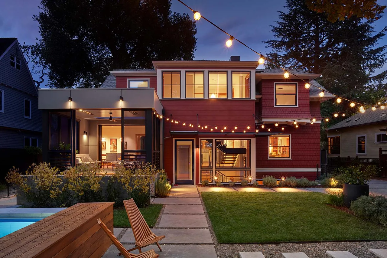 Two-story red house with shingle siding and a modern screened patio lit with string lights in the backyard at dusk.