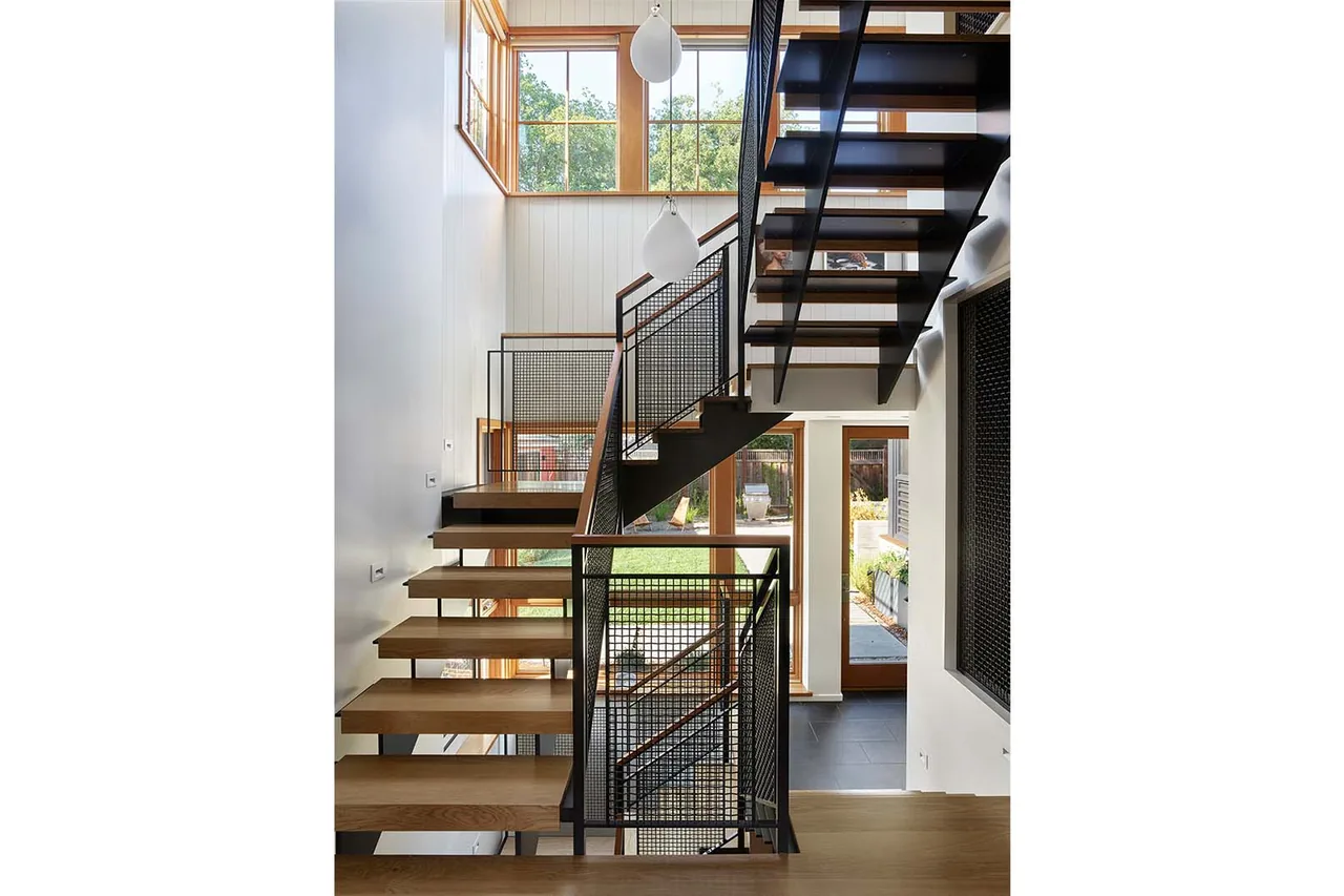 Modern staircase features open wood steps, black metal mesh railings, and large windows bringing in natural light.