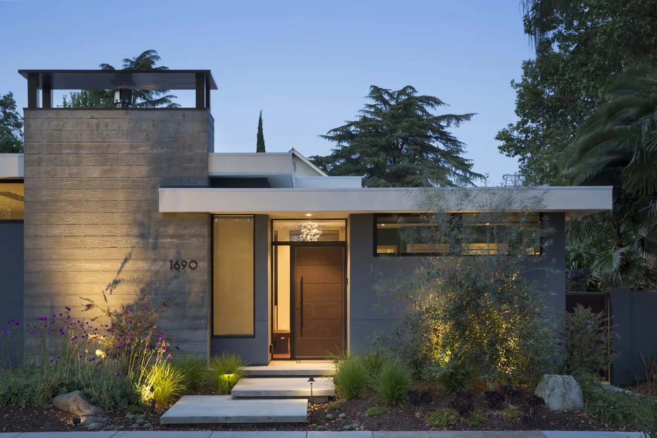 Modern house front exterior features a concrete chimney wall, flat roof, and dark framed transom windows above the entry door.