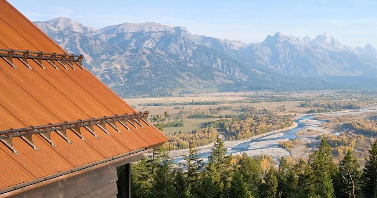 The photo shows a rust-colored standing seam metal roof of a mountain building overlooking a river valley and mountainous terrain.