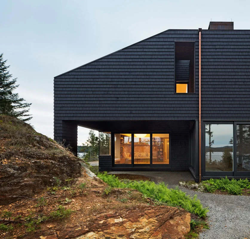 The Islesboro Residence features dark wood siding and large windows overlooking a waterfront setting in dusk light.