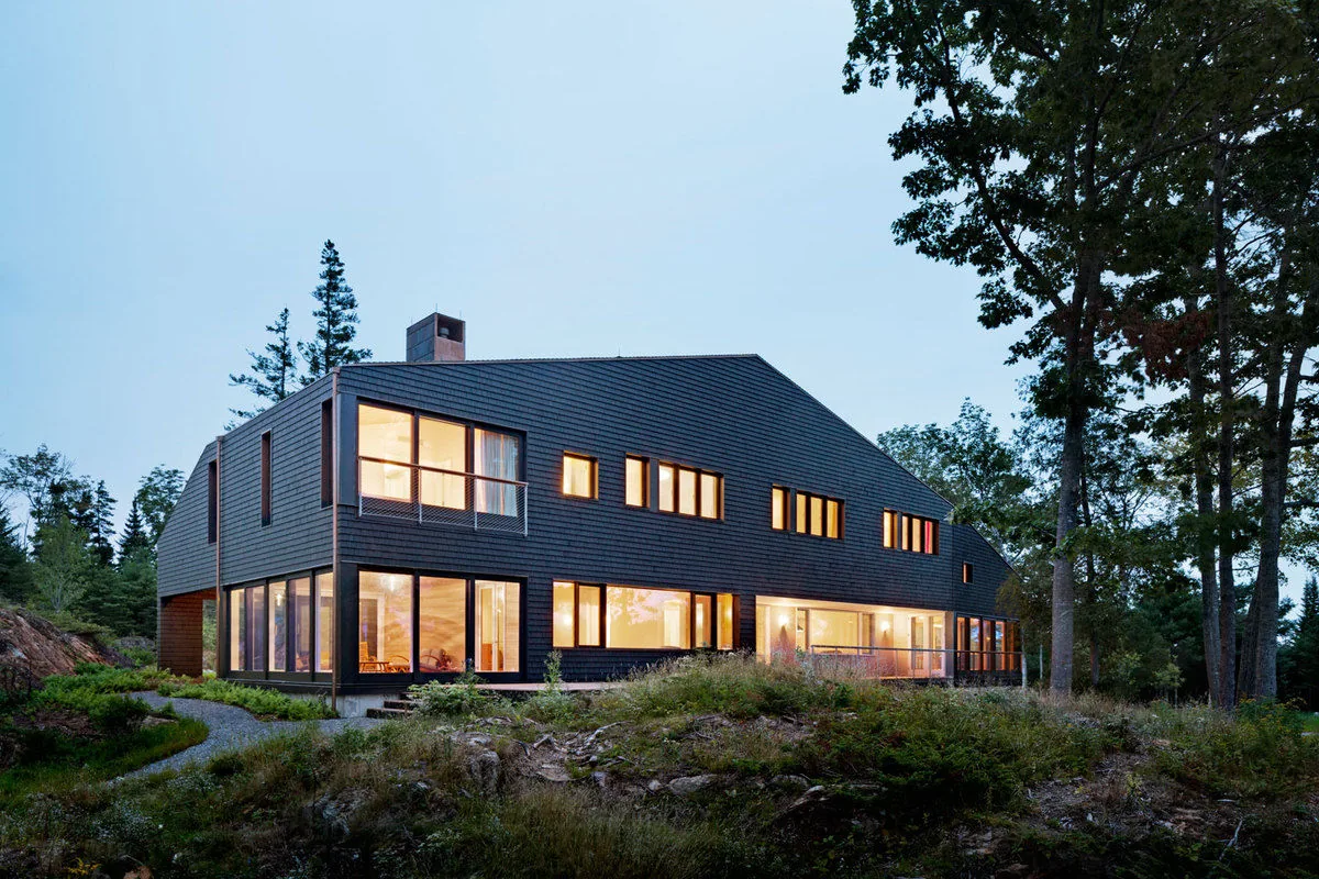 Islesboro Residence features black shingle siding with large windows and a gable roof, situated on rocky terrain at dusk.