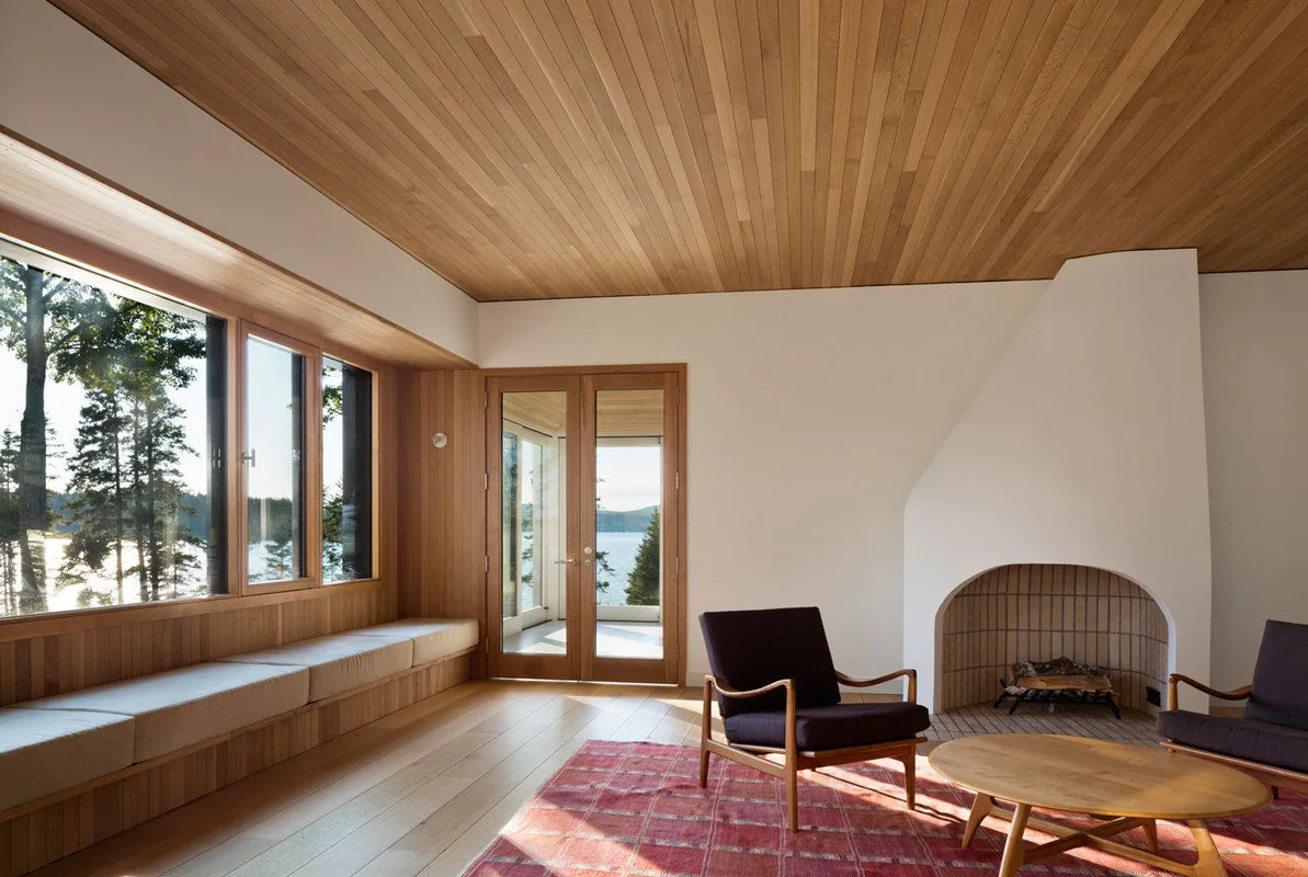 Living room with wood ceiling and wall paneling, a built-in bench seat by large windows, and a fireplace with a rounded opening.