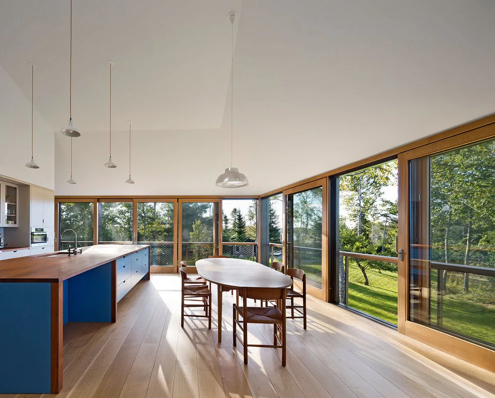 Dining area and kitchen with extensive floor-to-ceiling glass doors framed in wood, opening to a landscaped exterior.