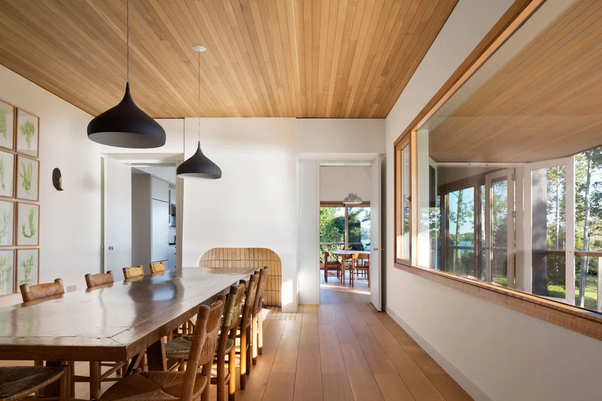 Islesboro Residence dining room with wood ceiling, long table, black pendant lights, and a large window to outdoor greenery.