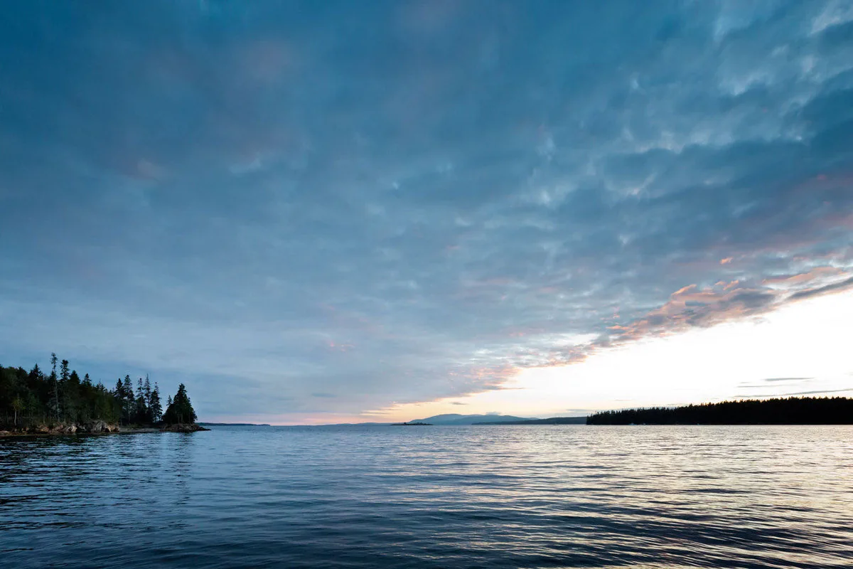 Waterfront landscape showing the calm sea with a cloudy sky and tree-lined shore at sunset.