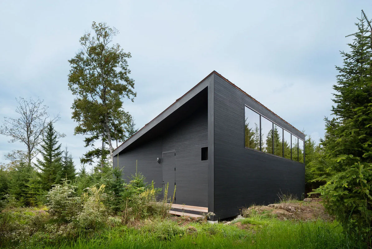 The Islesboro Boathouse and Studio features black painted horizontal wood siding and a raised foundation in a wooded setting.