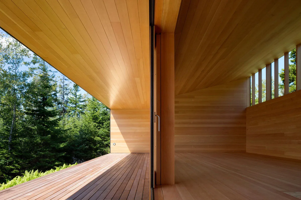 Wood-paneled room with matching ceiling and floor opens via sliding glass door to a covered deck overlooking dense forest.