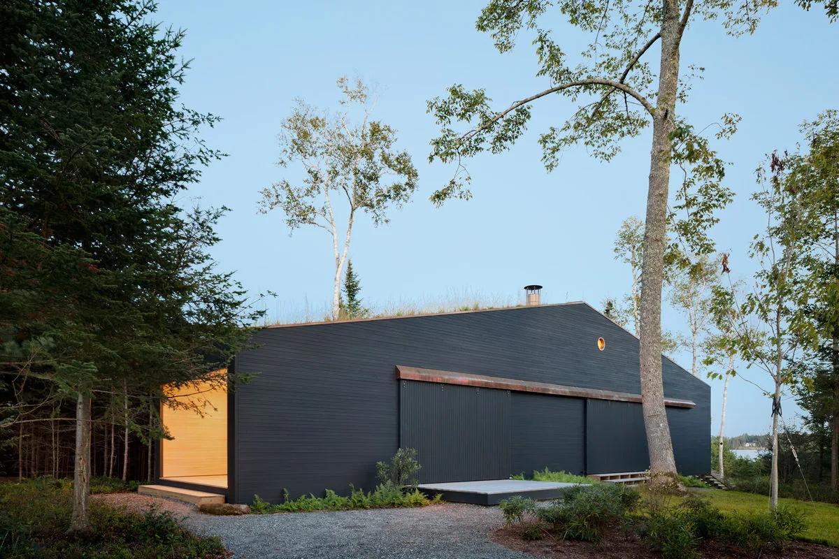 Islesboro Boathouse and Studio features black painted horizontal siding with large sliding doors, set against greenery and a clear sky.