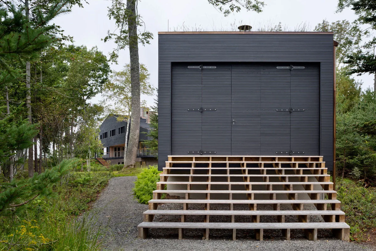 Small dark wood-sided boathouse with raised wooden steps and large doors in a wooded setting.