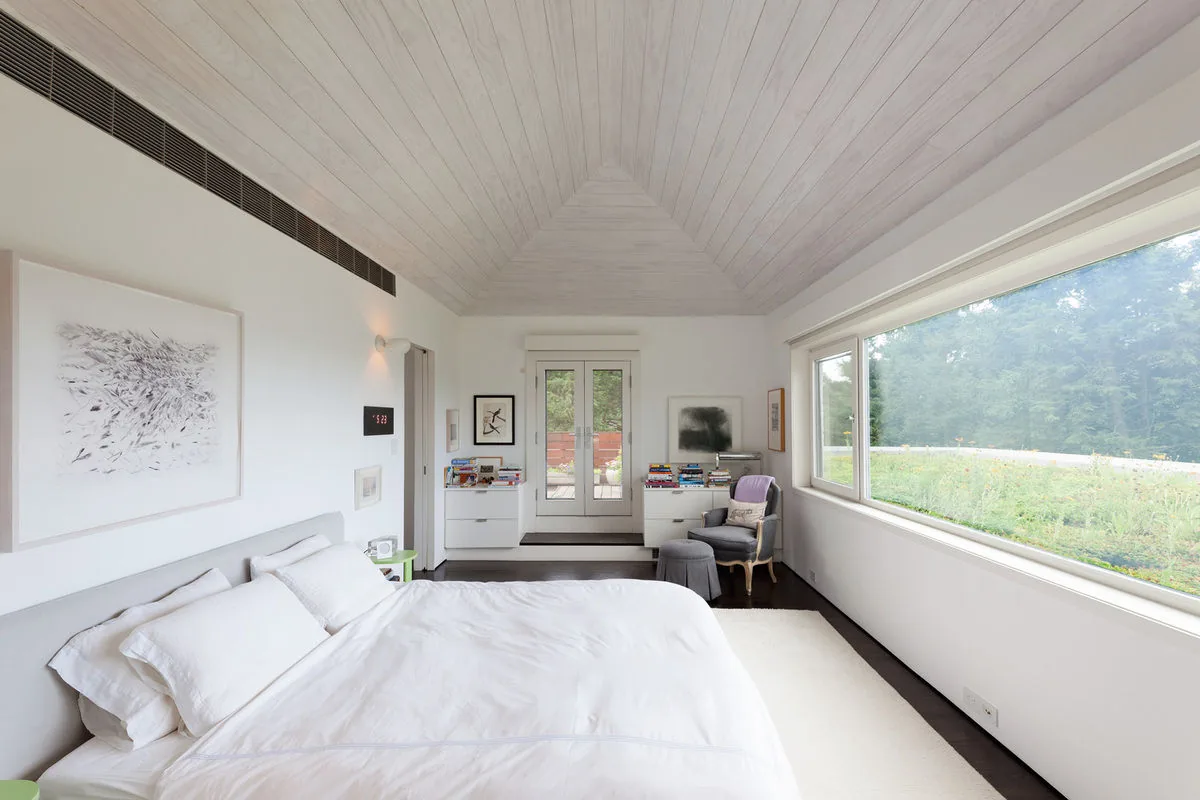 A minimalist bedroom features a vaulted ceiling, large horizontal window, and white walls in the Riverdale Residence.