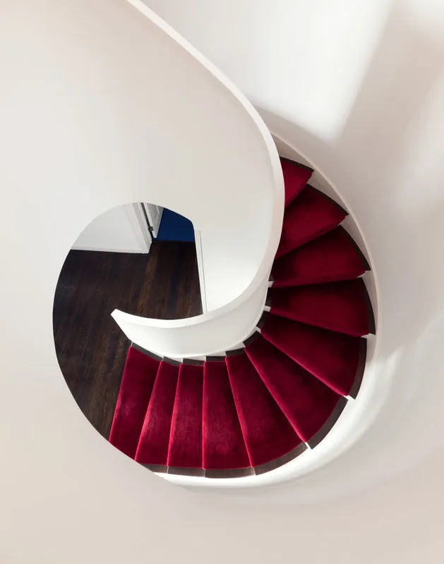Spiral staircase with red carpet treads and white walls inside the Riverdale Residence.