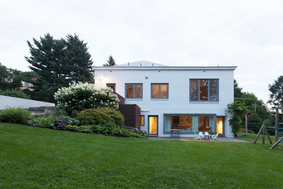 Rear view of the Riverdale Residence showing white brick walls, large windows, and a landscaped backyard patio area.