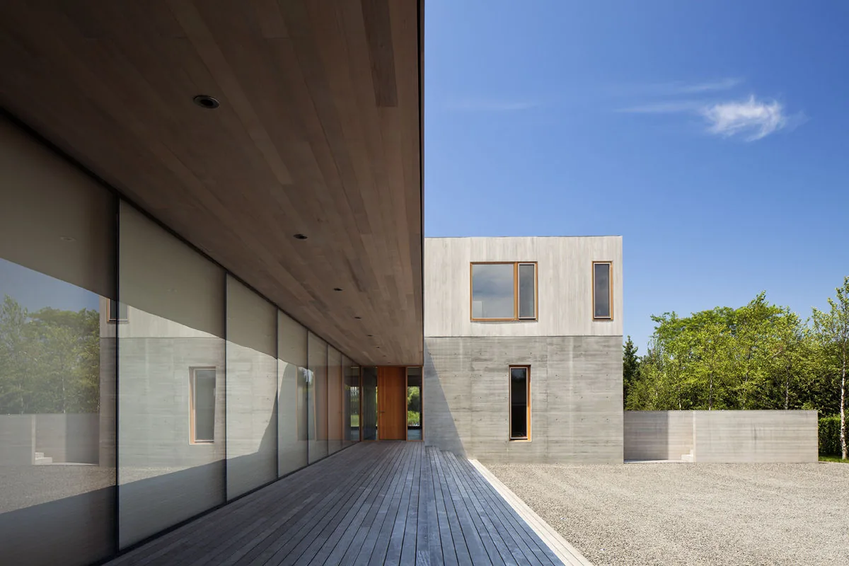 Covered wooden deck beside modern concrete and wood-clad two-story house with large glass windows.