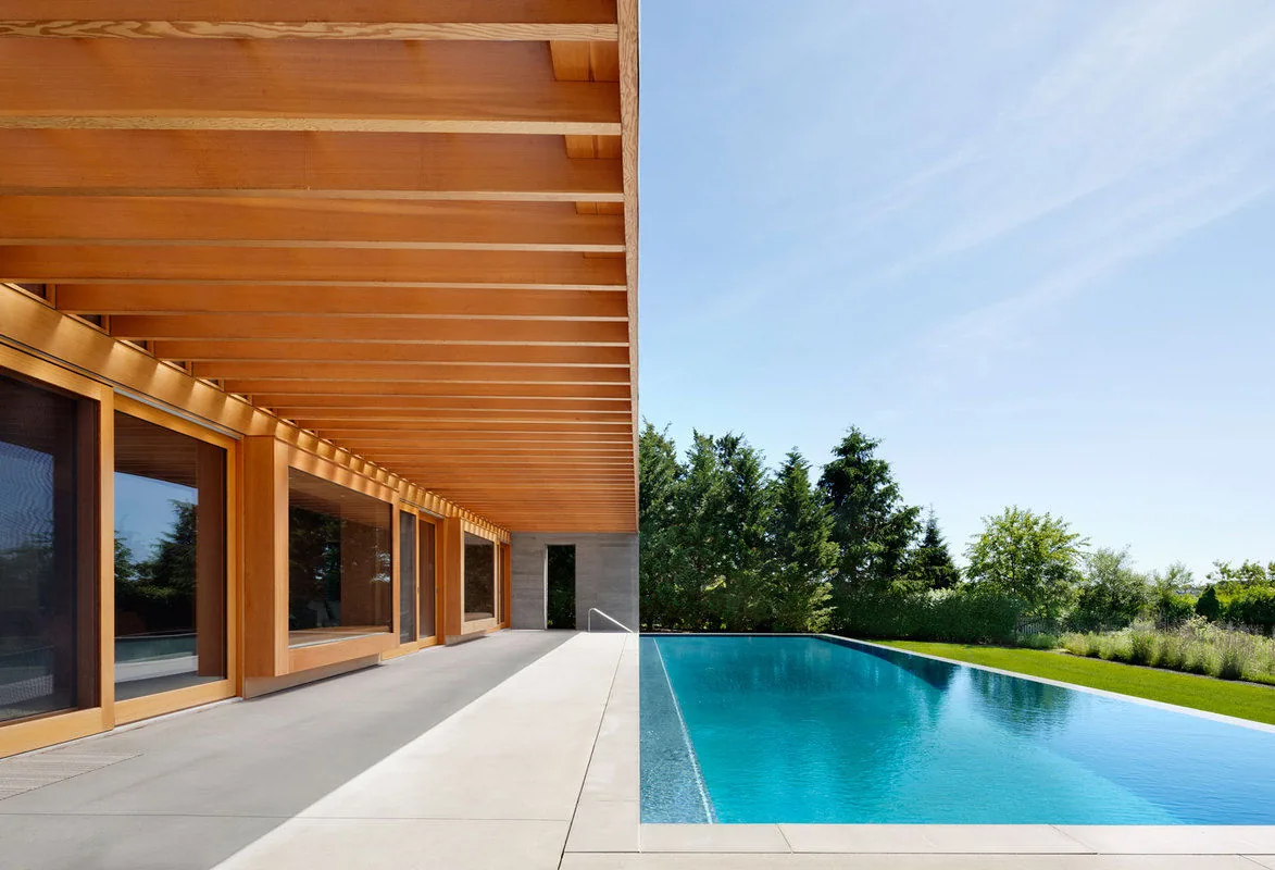 The Watermill Residence features a linear outdoor space with a wood-paneled pergola and a rectangular pool adjacent to the house.