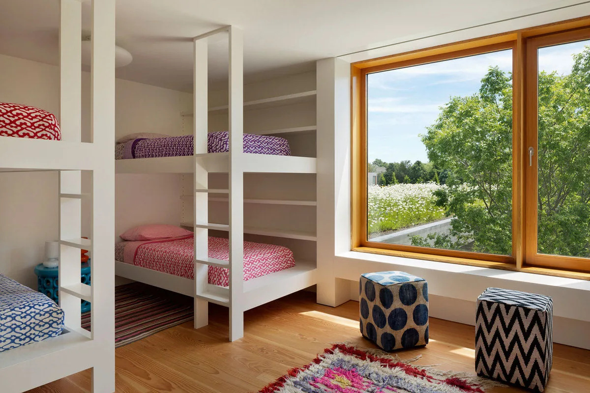 A bunk bed bedroom features white framing and a large wood-framed window with views of trees and flowers.