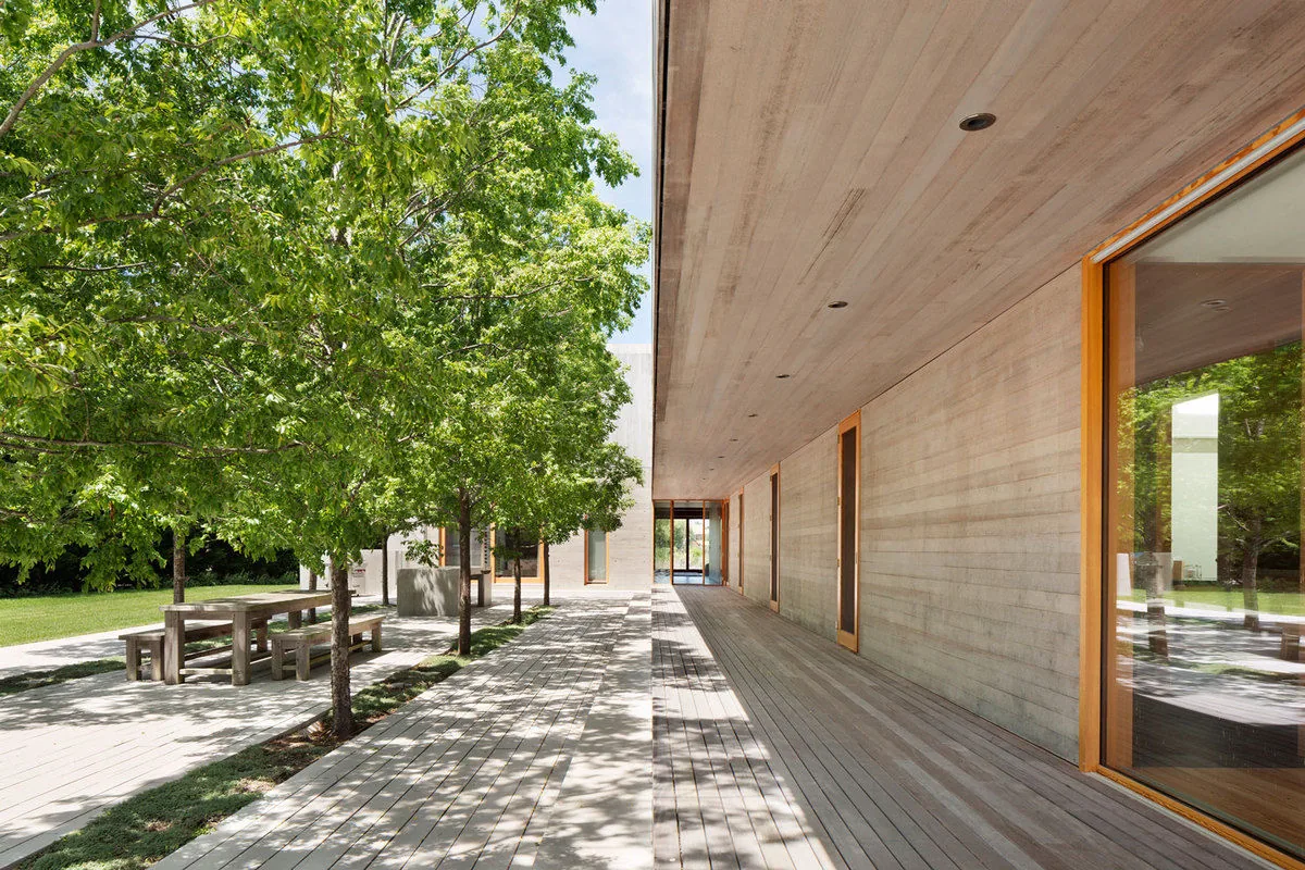 Board-formed concrete hallway opening to an outdoor seating area with trees and benches at Watermill Residence.