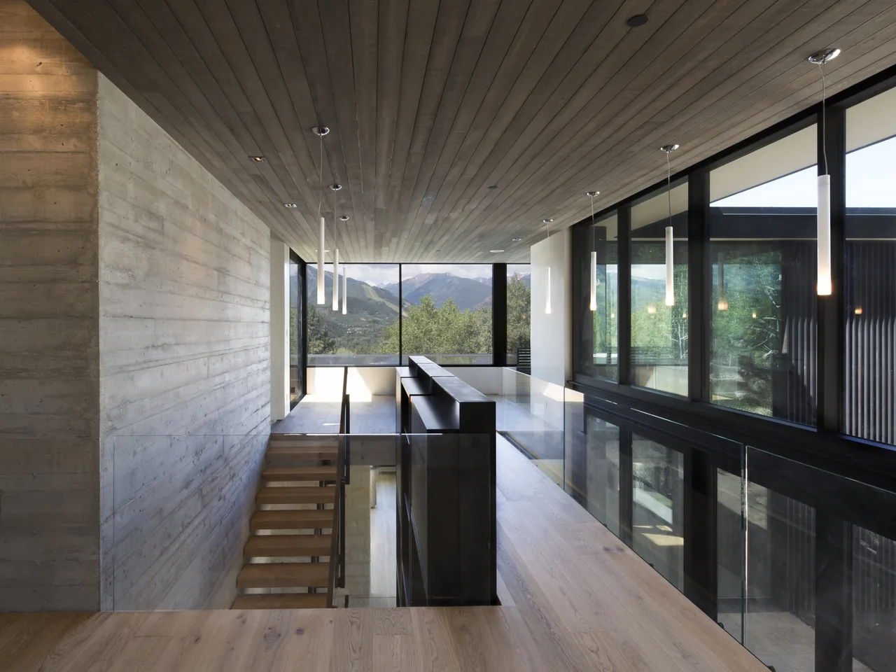 Hallway with wood ceiling and floor, concrete wall, and large black-framed windows overlooking mountain landscape.