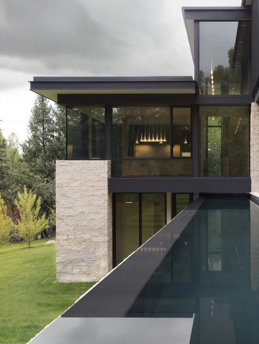 Exterior view of a modern residence featuring black window frames, stone masonry, and a reflecting pool along the side.