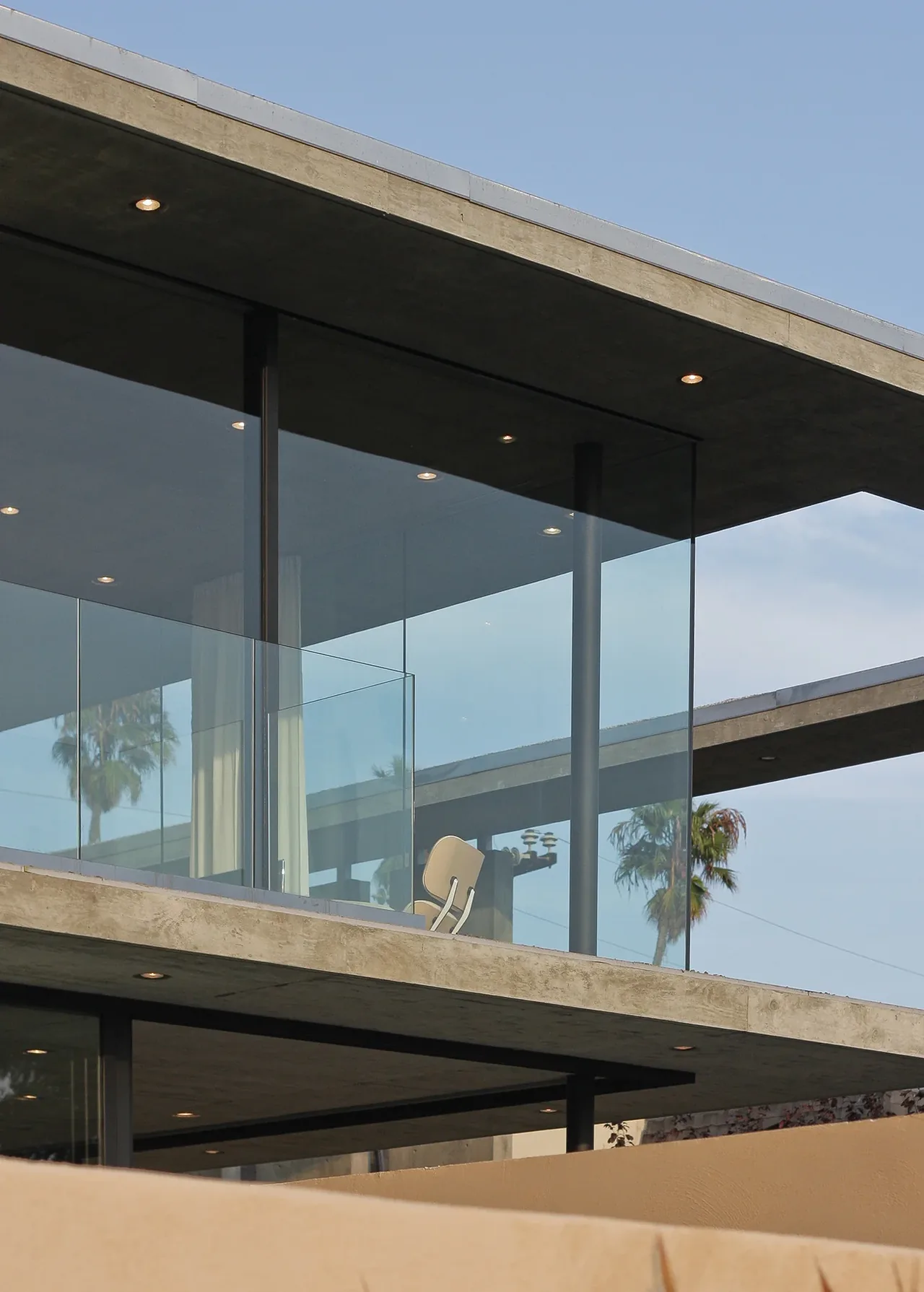 Modern concrete structure features floor-to-ceiling glazing and a glass balcony rail under a flat roof with recessed lighting.